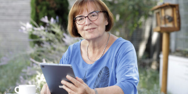 Frau sitzt mit Tablet im Garten an einem Tisch und lächelt in die Kamera