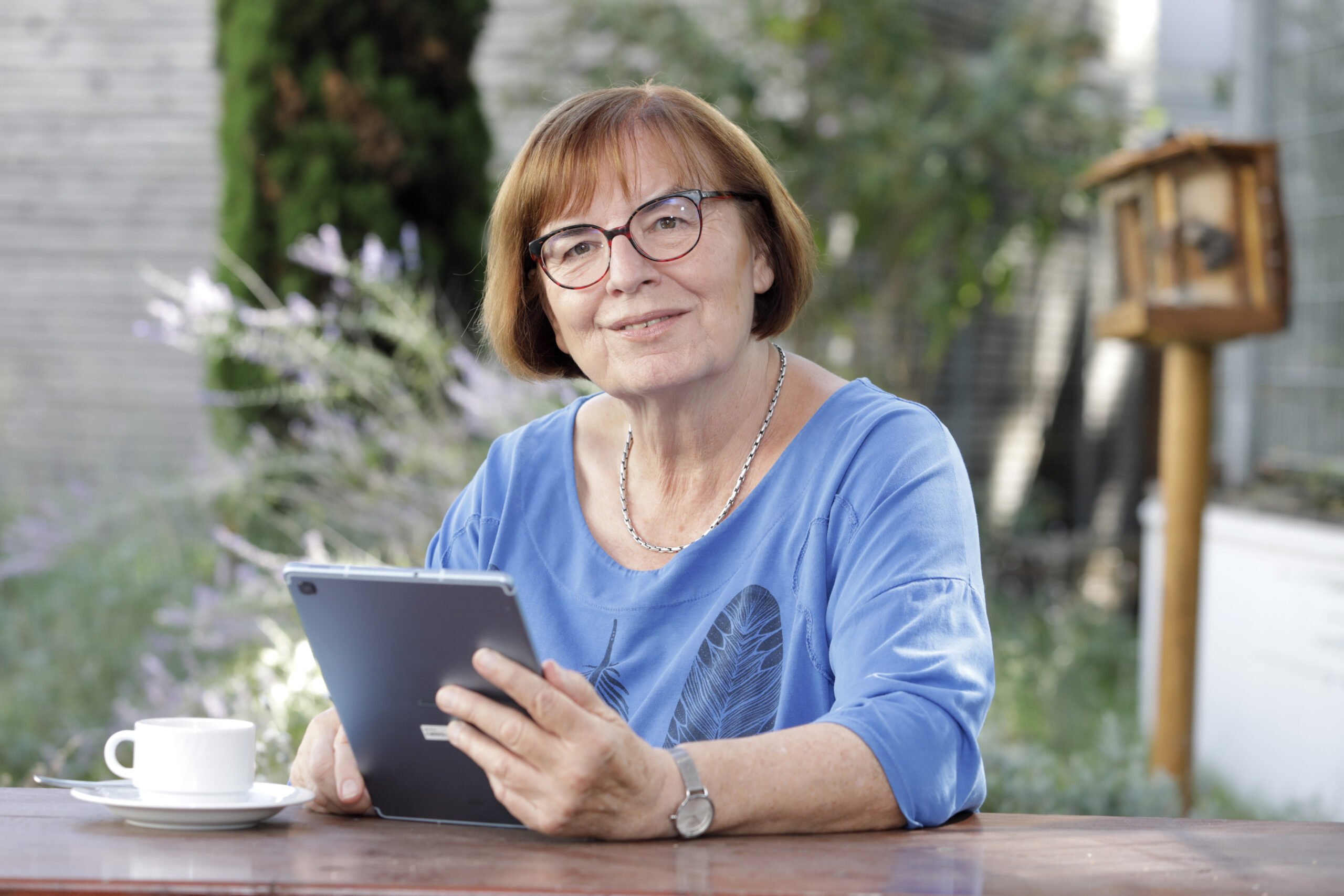 Frau sitzt mit Tablet im Garten an einem Tisch und lächelt in die Kamera