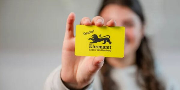 Eine Person hält eine gelbe Ehrenamtskarte Baden‑Württemberg mit schwarzem Löwenemblem und dem Wort „Danke!“ in die Kamera. Im Vordergrund ist die Karte scharf zu sehen, der Hintergrund ist unscharf.