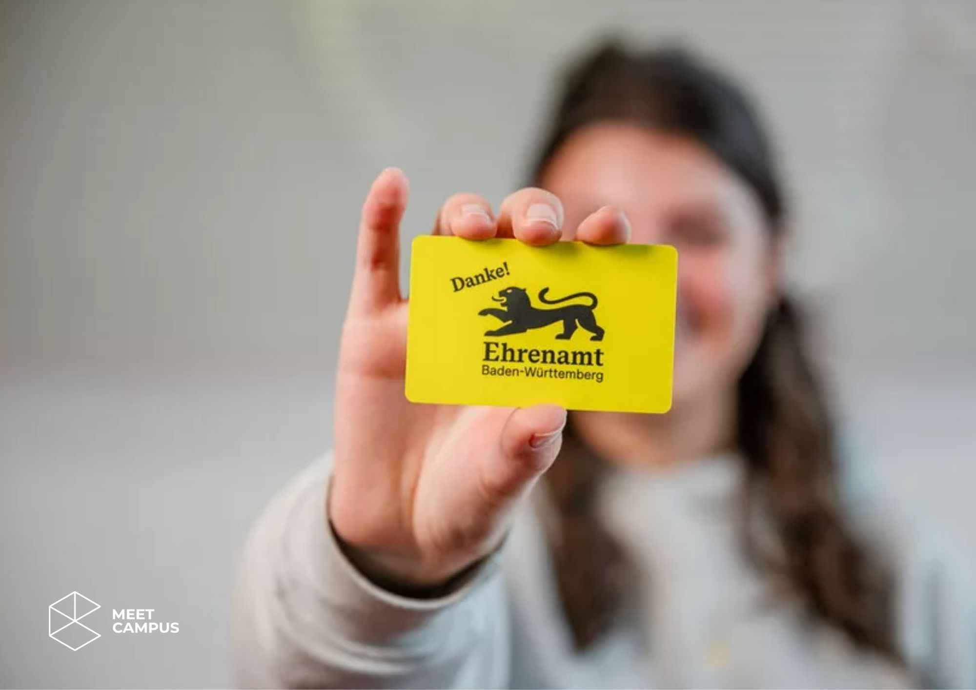 Eine Person hält eine gelbe Ehrenamtskarte Baden‑Württemberg mit schwarzem Löwenemblem und dem Wort „Danke!“ in die Kamera. Im Vordergrund ist die Karte scharf zu sehen, der Hintergrund ist unscharf.