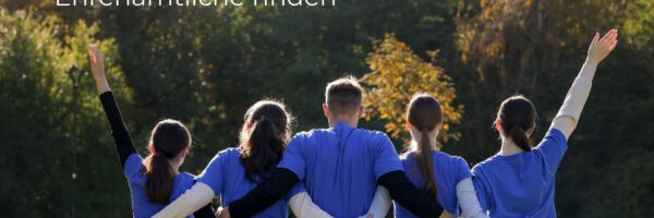 Gruppe von fünf Personen in blauen Shirts steht auf einer Wiese und schaut in Richtung Wald, mit erhobenen Armen; Text „Ehrenamtliche finden“ steht links im Bild.
