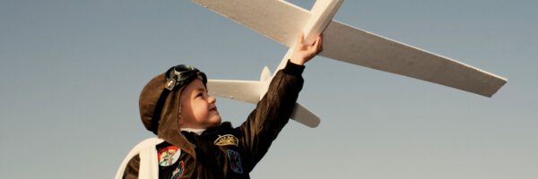 Ein Kind in einer Fliegerjacke und mit Pilotenkappe hält ein großes weißes Modellflugzeug hoch in den Himmel. Im Hintergrund ist ein klarer blauer Himmel zu sehen. Unten rechts steht das Wort ‚Zukunftsmut‘.