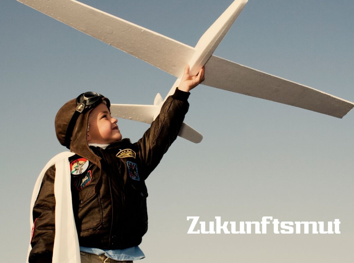 Ein Kind in einer Fliegerjacke und mit Pilotenkappe hält ein großes weißes Modellflugzeug hoch in den Himmel. Im Hintergrund ist ein klarer blauer Himmel zu sehen. Unten rechts steht das Wort ‚Zukunftsmut‘. Ein Kind in einer Fliegerjacke und mit Pilotenkappe hält ein großes weißes Modellflugzeug hoch in den Himmel. Im Hintergrund ist ein klarer blauer Himmel zu sehen. Unten rechts steht das Wort ‚Zukunftsmut‘.