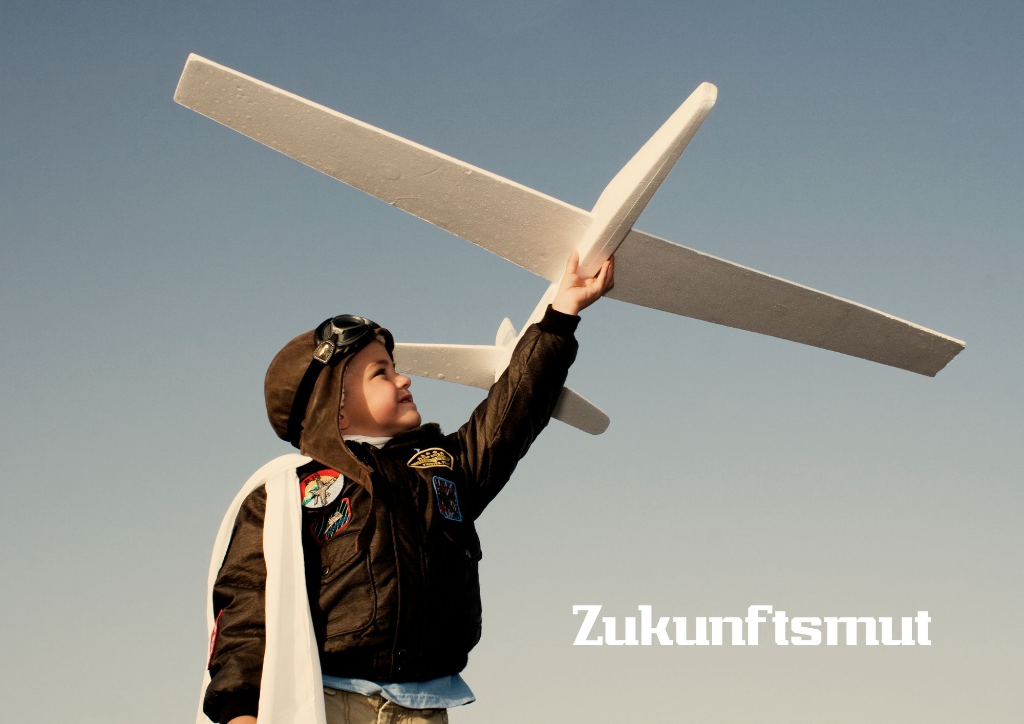 Ein Kind in einer Fliegerjacke und mit Pilotenkappe hält ein großes weißes Modellflugzeug hoch in den Himmel. Im Hintergrund ist ein klarer blauer Himmel zu sehen. Unten rechts steht das Wort ‚Zukunftsmut‘.
