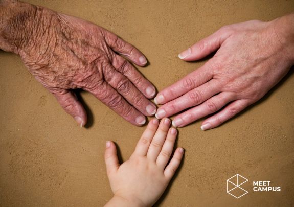 Drei Hände unterschiedlichen Alters liegen nebeneinander auf einer braunen Oberfläche: links eine ältere Hand, in der Mitte eine Kinderhand und rechts eine erwachsene Hand. Unten rechts befindet sich das MEET CAMPUS Logo mit geometrischem Design.