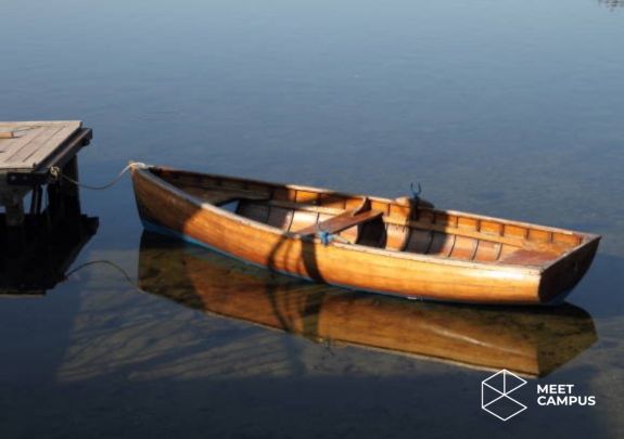 Ein hölzernes Ruderboot liegt an einem kleinen Steg auf klarem, ruhigem Wasser. Die Spiegelung des Boots ist im Wasser sichtbar. Unten rechts befindet sich das MEET CAMPUS Logo mit geometrischem Design.