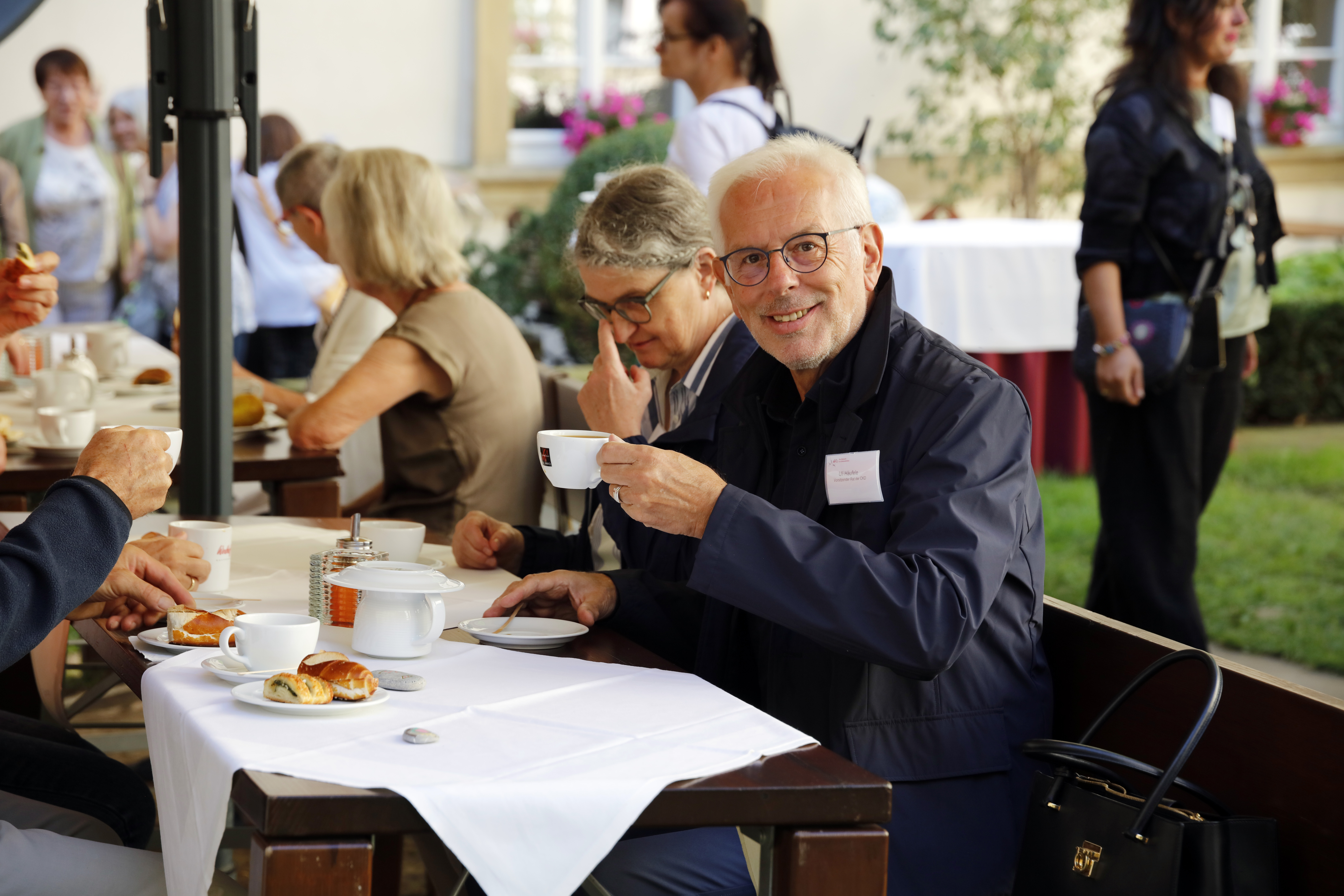 An einem Tisch im Freien sitzen mehrere Personen und trinken Kaffee und essen Kuchen. Ein Mann blickt freundlich in die Kamera und hebt seine Tasse.