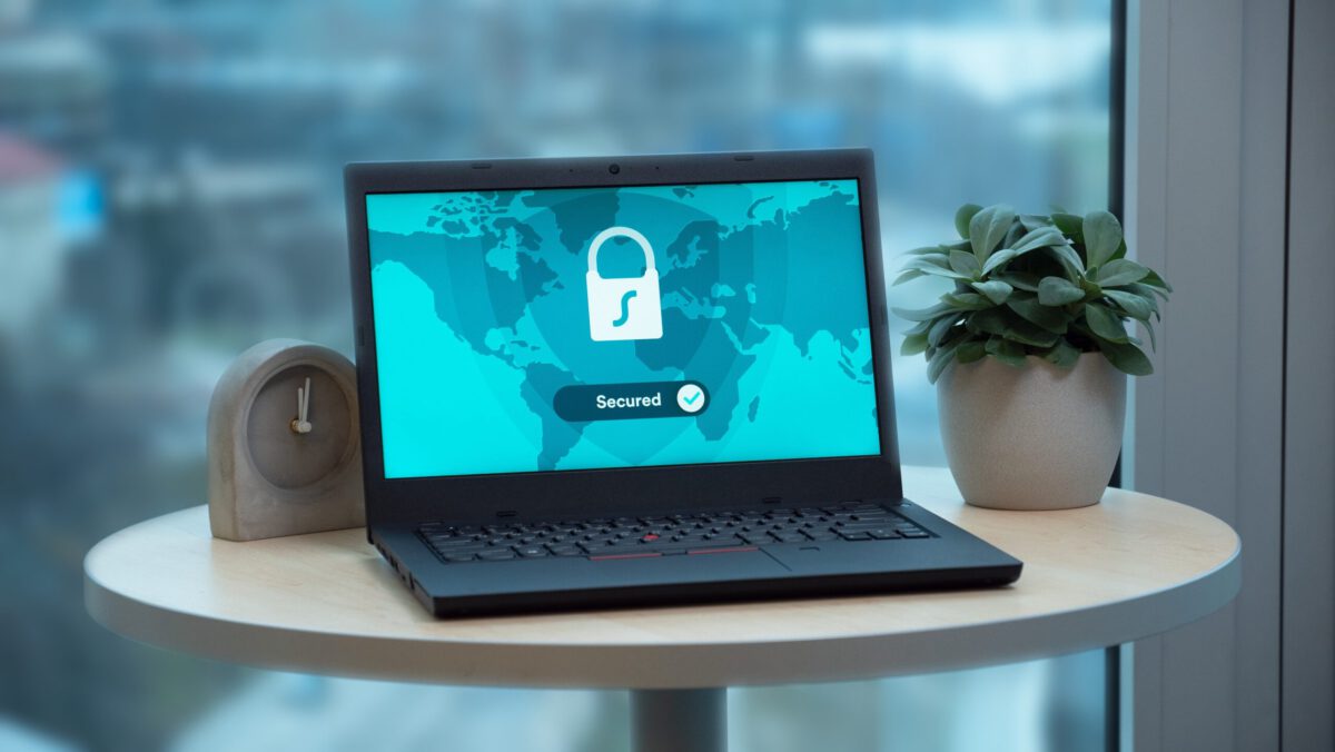 Laptop auf einem Tisch mit einem Sicherheitsschloss-Symbol und dem Wort 'Secured' auf dem Bildschirm, daneben eine Pflanze und eine Uhr, was auf ein Thema rund um Internetsicherheit oder Datenschutz hinweist. Hilfe für ein Digitales Ehrenamt.