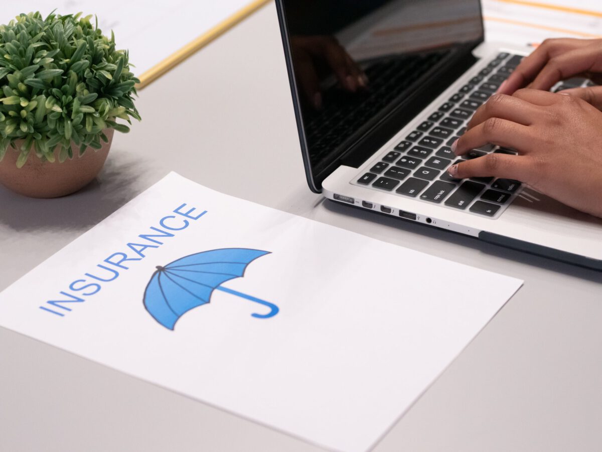 Hand tippt auf einem Laptop neben einem Blatt Papier mit der Aufschrift 'VERSICHERUNG' und einem blauen Regenschirm-Logo. Es geht um Versicherungen im Ehrenamt.