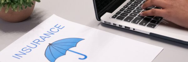 Hand tippt auf einem Laptop neben einem Blatt Papier mit der Aufschrift 'VERSICHERUNG' und einem blauen Regenschirm-Logo. Es geht um Versicherungen im Ehrenamt.